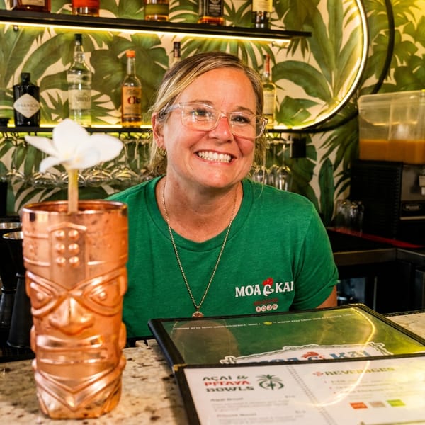 Smiling woman in a green Moa Kai shirt behind a bar with tropical wallpaper and drinks.