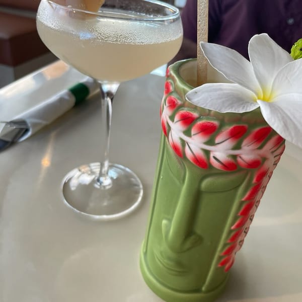 Two cocktails on a table, one in a coupe glass with lychees, the other in a green tiki mug.