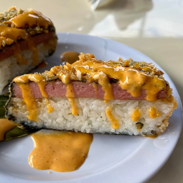 A slice of Spam musubi with rice, Spam, and a drizzle of orange sauce on a white plate.