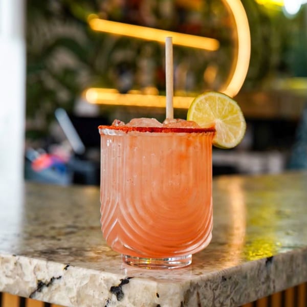 Pink cocktail with a chili-lime rim, lime wedge, and straw on a granite bar.