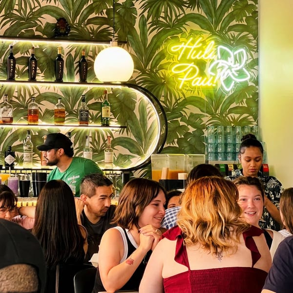 People enjoying drinks and conversation at a lively bar with tropical leaf wallpaper and a neon sign.