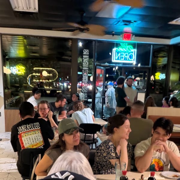Diners seated at tables and a counter in a bustling restaurant with neon signs visible through windows.